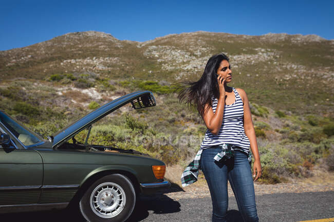 Mujer de raza mixta hablando con un teléfono inteligente de pie en la carretera junto a un coche averiado con capó abierto. viaje de verano por carretera en un camino rural junto a la costa. - foto de stock