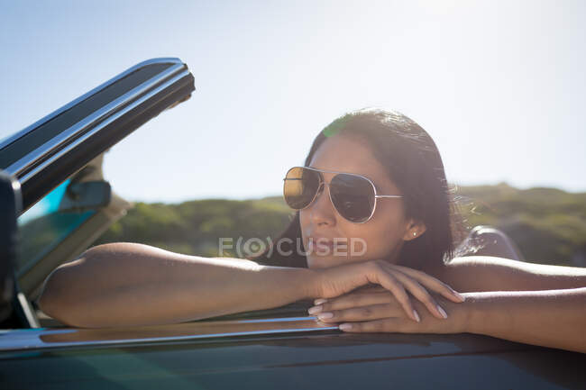 Mixed race woman on sunny day sitting in convertible car leaning on the car doors. summer road trip on a country highway by the coast. — Stock Photo