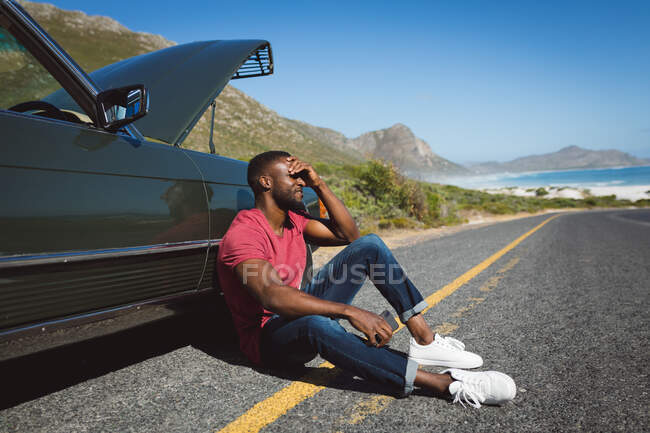 African american man holding smartphone sitting on road beside broken-down car with open bonnet. Summer road trip on a country road by the coast. — Stock Photo