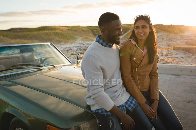 Casal diverso sentado em um carro conversível olhando um para o outro e sorrindo. viagem de estrada de verão em uma estrada rural pela costa. — Fotografia de Stock