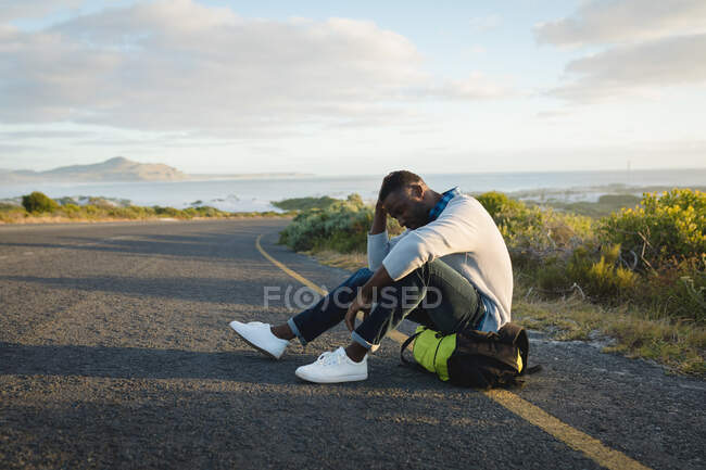 Afroamerikaner sitzt auf seinem Rucksack am Straßenrand. Sommerreisen auf der Landstraße an der Küste. — Stockfoto