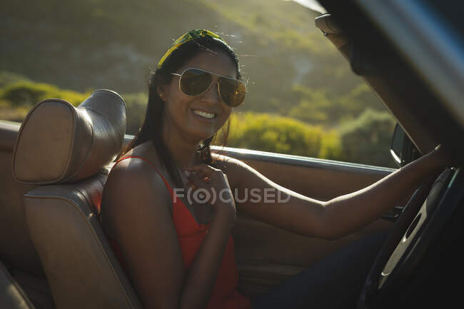 Mujer de raza mixta conduciendo en un día soleado en un coche descapotable sosteniendo el volante y sonriendo. Viaje de verano por carretera en una carretera rural junto a la costa. - foto de stock
