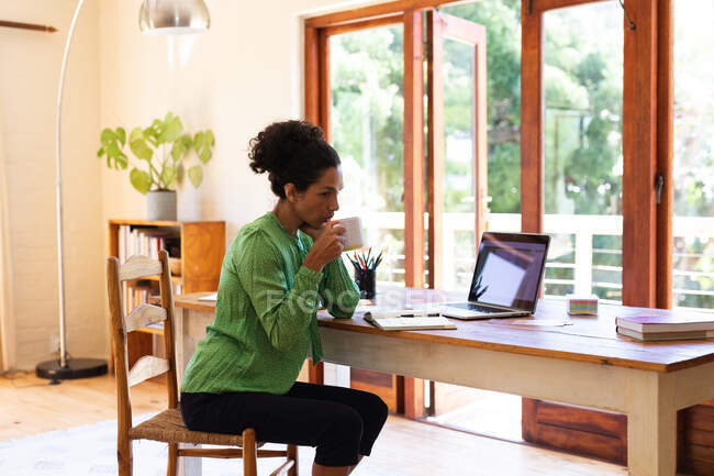 Белая женщина пьет кофе за столом, работает из дома. Оставаться дома в изоляции во время карантинной изоляции. — стоковое фото