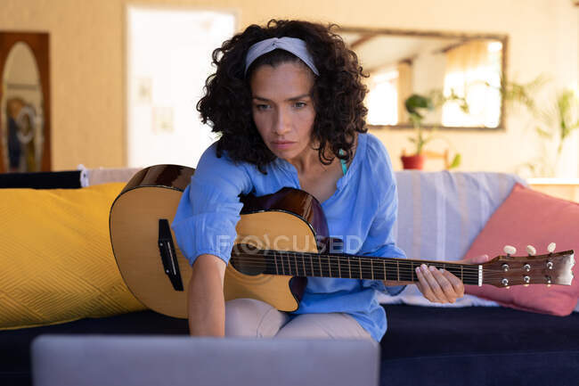 Caucasian woman holding guitar using laptop sitting on sofa at home. Staying at home in self isolation during quarantine lockdown. — Stock Photo