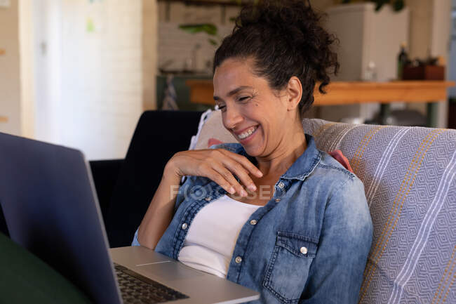 Mujer caucásica sonriendo con el ordenador portátil en la videollamada sentado en el sofá en casa. Permanecer en casa en aislamiento durante el bloqueo de cuarentena. - foto de stock