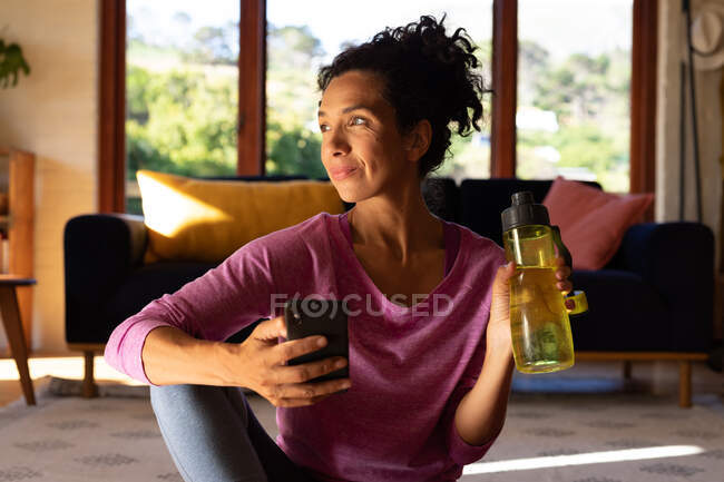 Mujer caucásica sosteniendo botella de agua y teléfono inteligente tomando un descanso de hacer ejercicio en casa. Permanecer en casa en aislamiento durante el bloqueo de cuarentena. - foto de stock