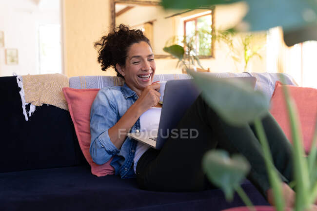Mujer caucásica sonriendo con el ordenador portátil en la videollamada sentado en el sofá en casa. Permanecer en casa en aislamiento durante el bloqueo de cuarentena. - foto de stock