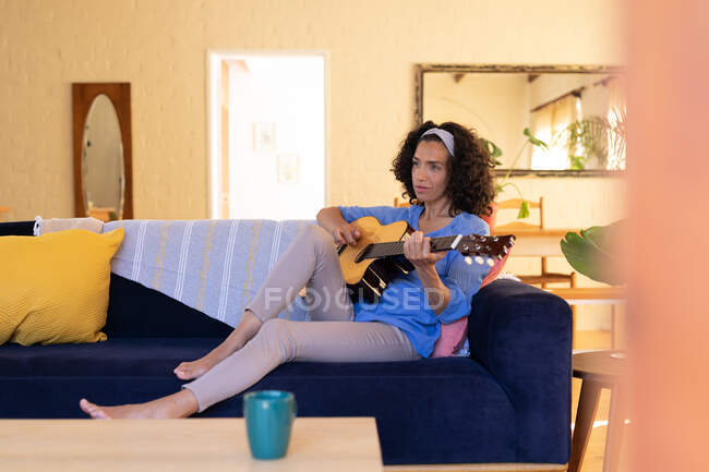 Белая женщина, играющая на гитаре, сидит дома на диване. Оставаться дома в изоляции во время карантинной изоляции. — стоковое фото