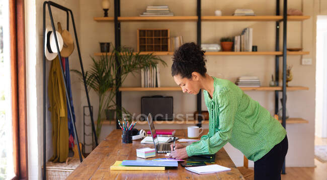 Mujer caucásica escribiendo, de pie junto al escritorio, trabajando desde casa. Permanecer en casa en aislamiento durante el bloqueo de cuarentena. - foto de stock