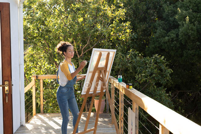 Mulher branca pintando quadro em pé no terraço ensolarado. Ficar em casa em auto-isolamento durante o bloqueio de quarentena. — Fotografia de Stock