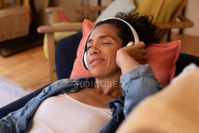 Mujer caucásica acostada en un sofá con auriculares puestos, relajándose en casa. Permanecer en casa en aislamiento durante el bloqueo de cuarentena. - foto de stock