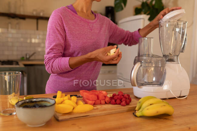 Parte central da mulher colocando frutas no espremedor na cozinha em casa. Ficar em casa em auto-isolamento durante o bloqueio de quarentena. — Fotografia de Stock