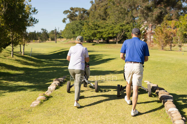 Кавказький старший чоловік і жінка ідуть через поле для гольфу з мішками для гольфу. Хобі для гольфу, здоровий спосіб життя на пенсії — стокове фото