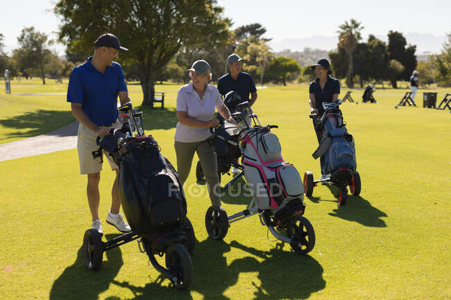 Quatro homens e mulheres caucasianos a atravessar o campo de golfe com sacos de golfe. Esportes de golfe passatempo, estilo de vida de aposentadoria saudável — Fotografia de Stock