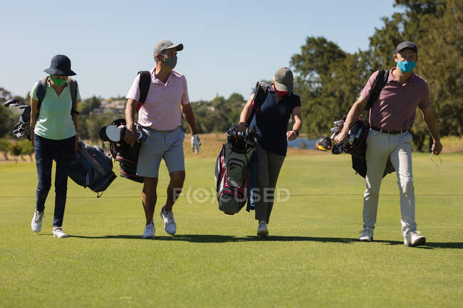 Cuatro hombres y mujeres mayores caucásicos con máscaras faciales caminando por el campo de golf sosteniendo bolsas de golf. deporte de golf hobby, estilo de vida de jubilación saludable durante coronavirus covid 19 pandemia. - foto de stock
