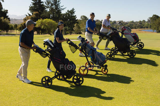 Quatre hommes et femmes âgés caucasiens marchant à travers le terrain de golf tenant des sacs de golf. Golf passe-temps sportif, mode de vie sain à la retraite — Photo de stock