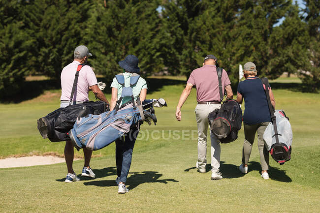 Quatro homens e mulheres caucasianos a atravessar o campo de golfe com sacos de golfe. passatempo esportivo de golfe, estilo de vida de aposentadoria saudável durante coronavírus covid 19 pandemia. — Fotografia de Stock