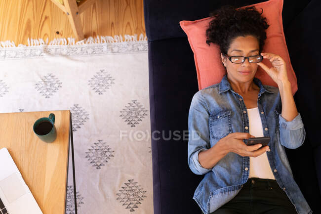 Mujer caucásica tumbada en el sofá con la celebración de teléfono inteligente, relajarse en casa. Permanecer en casa en aislamiento durante el bloqueo de cuarentena. - foto de stock