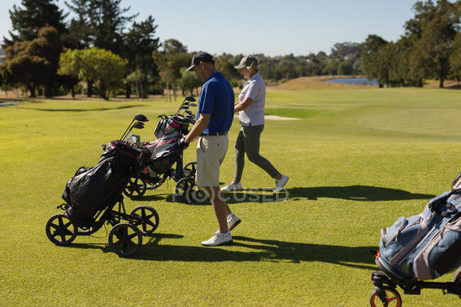 Blanc senior homme et femme marchant à travers le terrain de golf tenant des sacs de golf. golf passe-temps sportif, mode de vie sain à la retraite — Photo de stock