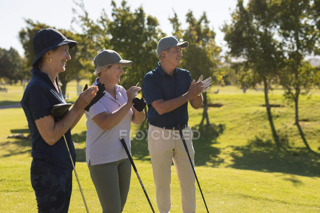 Tres ancianos caucásicos sosteniendo palos de golf y aplaudiendo. deportes de golf hobby, estilo de vida de jubilación saludable - foto de stock