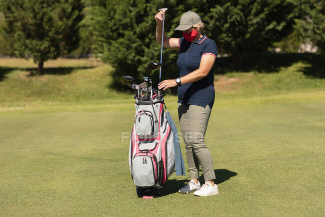 Белая женщина в маске для лица достает клюшку для гольфа из сумки для гольфа. гольф спортивное хобби, здоровый пенсионный образ жизни во время коронавируса ковид 19 пандемии. — стоковое фото