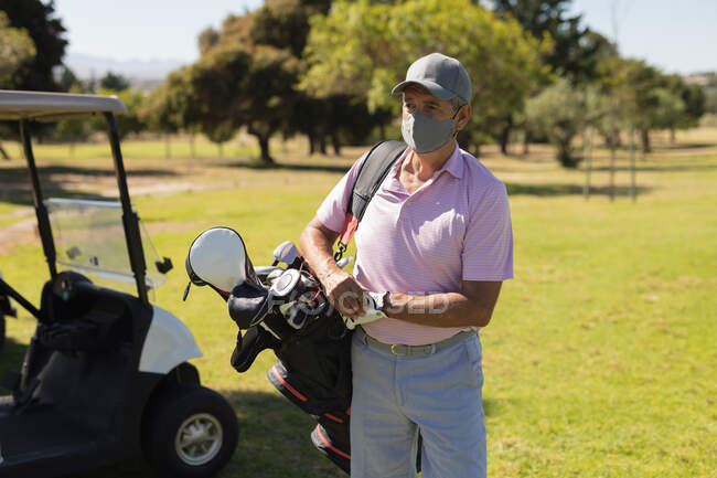 Uomo anziano caucasico con maschera facciale che cammina attraverso il campo da golf tenendo borsa da golf. Golf sport hobby, sano stile di vita di pensionamento durante coronavirus covid 19 pandemia. — Foto stock