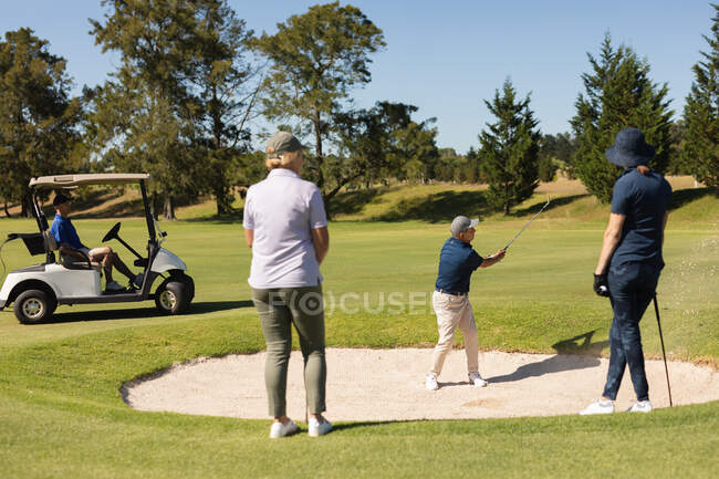 Дві кавказькі жінки старшого віку спостерігають, як чоловік стріляє в бункер. гольф спортивне хобі, здоровий спосіб життя на пенсії . — стокове фото