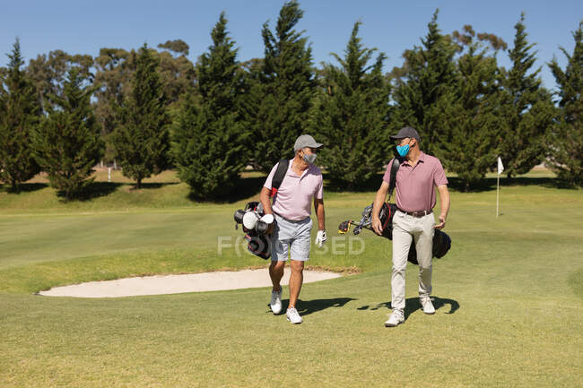 Dois homens caucasianos com máscaras faciais a atravessar o campo de golfe com sacos de golfe. passatempo esportivo de golfe, estilo de vida de aposentadoria saudável durante coronavírus covid 19 pandemia. — Fotografia de Stock