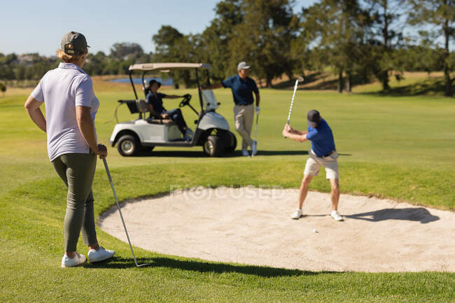 Cuatro hombres y mujeres mayores caucásicos jugando al golf viendo a un hombre preparando un tiro desde el búnker. Golf deportes hobby, estilo de vida de jubilación saludable. - foto de stock