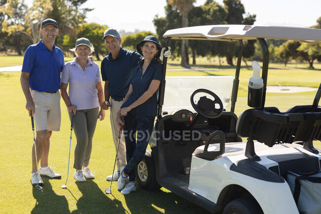 Cuatro hombres y mujeres mayores caucásicos de pie junto al buggy de golf mirando a la cámara y sonriendo. deportes de golf hobby, estilo de vida de jubilación saludable - foto de stock