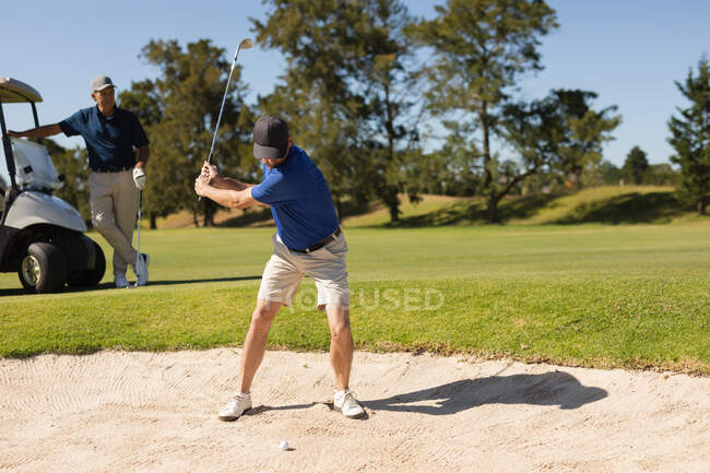 Hombre mayor caucásico viendo a otro hombre sosteniendo el club de golf preparándose para disparar en el búnker. deportes de golf hobby, estilo de vida de jubilación saludable. - foto de stock
