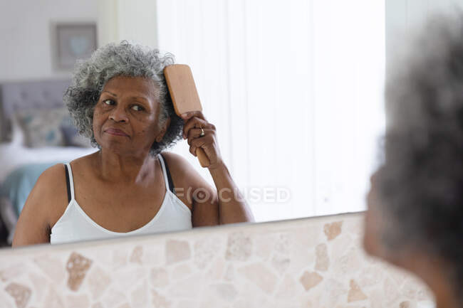 Pensativo afroamericano anciano mujer cepillarse el pelo mientras se mira en el espejo en casa. permaneciendo en casa en aislamiento en cuarentena - foto de stock