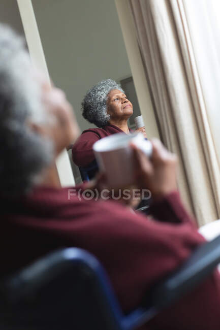 Mulher sênior afro-americana atenciosa segurando xícara de café olhando pela janela enquanto sentada em cadeira de rodas em casa. ficar em casa em auto-isolamento em quarentena — Fotografia de Stock