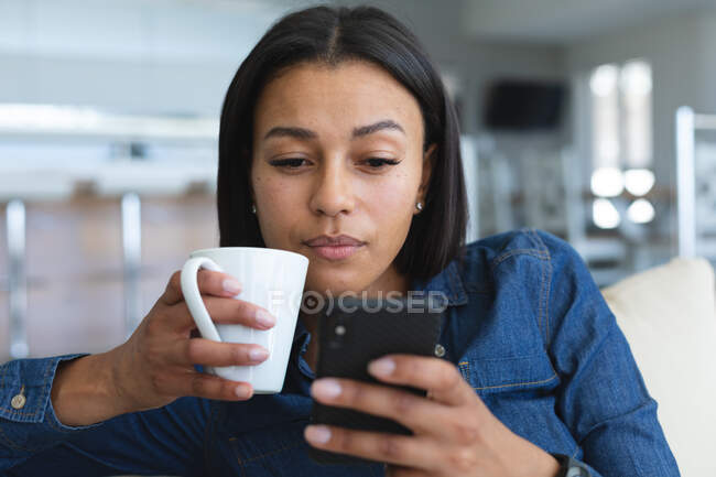 Mujer afroamericana sosteniendo una taza de café usando un teléfono inteligente mientras está sentada en el sofá de su casa. permaneciendo en casa en aislamiento en cuarentena - foto de stock