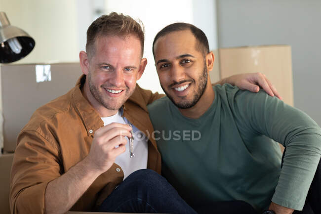 Casal masculino gay multi étnico sentado cercado por caixas olhando para a câmera sorrindo em casa. Aproveitando o tempo em casa em auto-isolamento durante o bloqueio de quarentena. — Fotografia de Stock