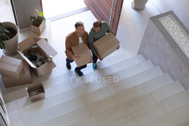 Couple homosexuel multi ethnique tenant des boîtes marchant dans les escaliers à la maison. Profiter du temps passé à la maison en isolement personnel pendant le confinement en quarantaine. — Photo de stock