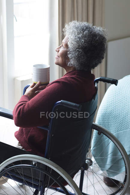 Donna anziana afroamericana con una tazza di caffè sorridente mentre guarda fuori dalla finestra mentre è seduta sulla sedia a rotelle a casa. stare a casa in isolamento personale in quarantena — Foto stock