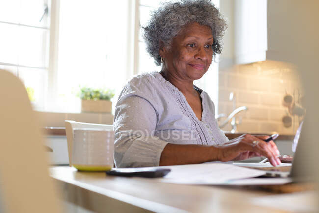 Mujer mayor afroamericana sonriendo mientras usa el portátil en casa. permaneciendo en casa en aislamiento en cuarentena - foto de stock