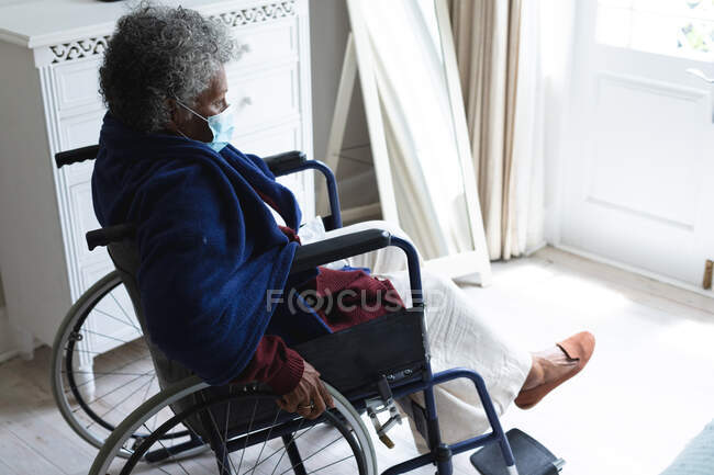 Femme âgée afro-américaine réfléchie portant un masque assis sur un fauteuil roulant à la maison. rester à la maison en isolement personnel en quarantaine — Photo de stock