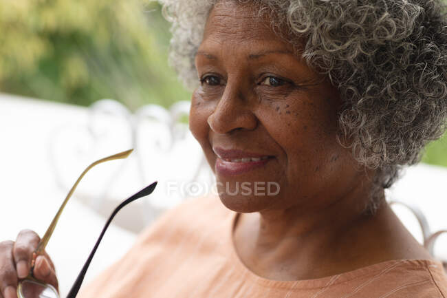 Retrato da mulher afro-americana idosa segurando óculos sorrindo enquanto estava sentada no alpendre. permanecer em auto-isolamento em quarentena — Fotografia de Stock
