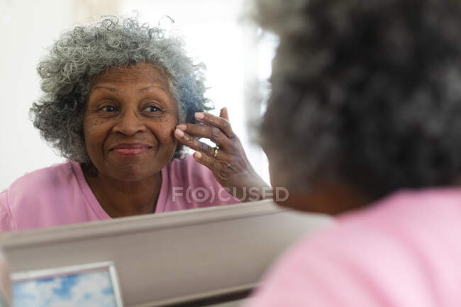 Африканская старшая женщина улыбается, когда трогает лицо, глядя в зеркало дома. оставаться дома в изоляции в карантинной изоляции — стоковое фото