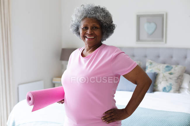 Portrait de femme afro-américaine âgée souriant tout en tenant tapis de yoga à la maison. rester à la maison en isolement personnel en quarantaine — Photo de stock