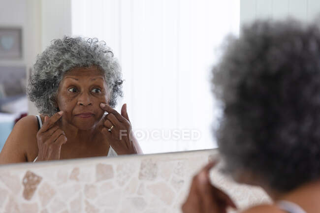 Eine afroamerikanische Seniorin berührt ihr Gesicht, während sie zu Hause in den Spiegel schaut. Zu Hause bleiben in Selbstisolierung in Quarantäne — Stockfoto