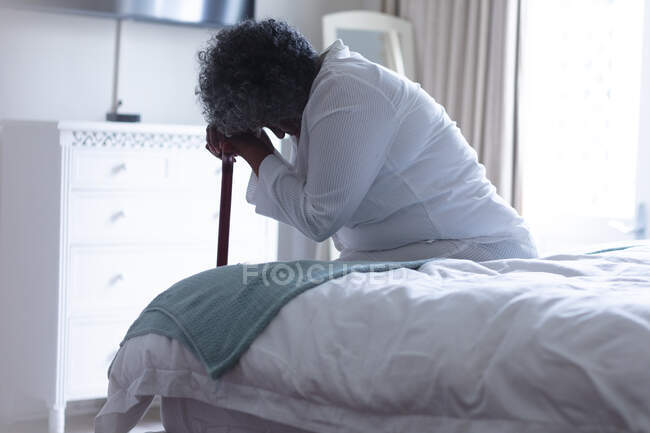 Thoughtful african american senior woman holding walking stick while sitting on bed at home. staying at home in self isolation in quarantine lockdown — Stock Photo