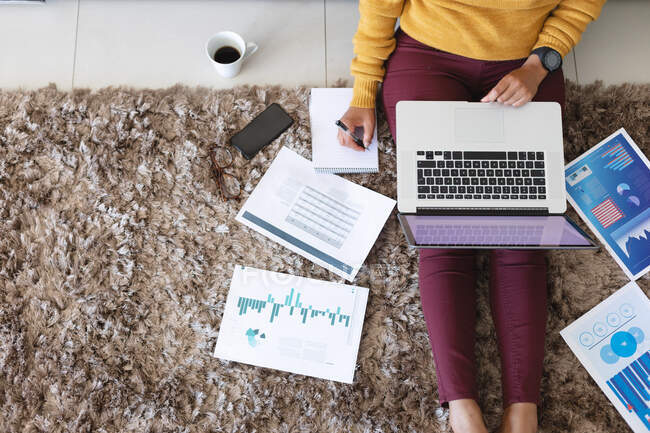 Überkopfansicht einer afrikanisch-amerikanischen Frau mit Laptop, die Notizen macht, während sie von zu Hause aus arbeitet. Zu Hause bleiben in Selbstisolierung in Quarantäne — Stockfoto