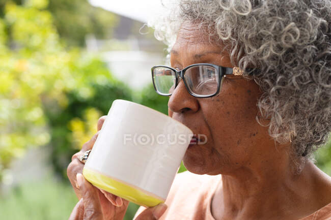 Закрыть вдумчивую африканскую старшую женщину, держащую чашку кофе, сидя на крыльце. оставаться в изоляции в карантинной изоляции — стоковое фото
