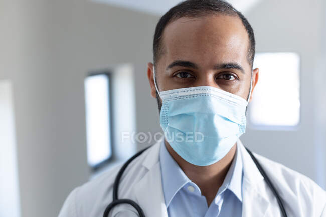 Ritratto di medico maschio di razza mista che indossa una maschera che guarda la macchina fotografica. operatore medico professionista che indossa stetoscopio e camice da laboratorio. — Foto stock