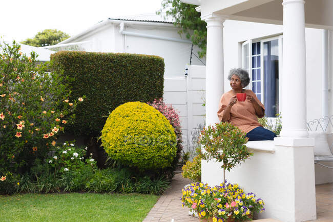 Mujer mayor afroamericana reflexiva sosteniendo una taza de café mientras está sentada en el porche de la casa. permaneciendo en aislamiento en cuarentena - foto de stock