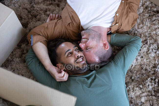 Casal masculino gay multi étnico deitado no chão e abraçando em casa. aproveitando o tempo em casa em auto-isolamento durante o bloqueio de quarentena. — Fotografia de Stock