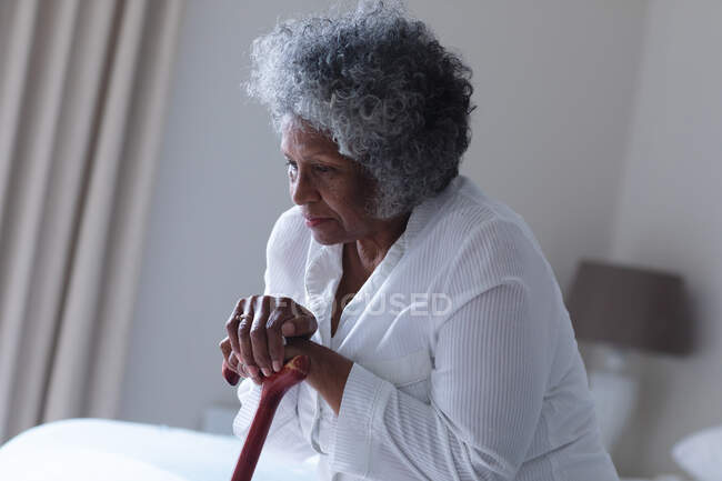 Mujer mayor afroamericana reflexiva sosteniendo bastón mientras está sentada en la cama en casa. permaneciendo en casa en aislamiento en cuarentena - foto de stock